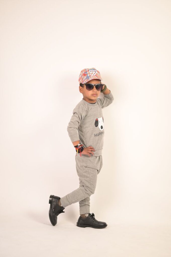A fashionable young boy wearing sunglasses and a cap poses confidently in a gray outfit.